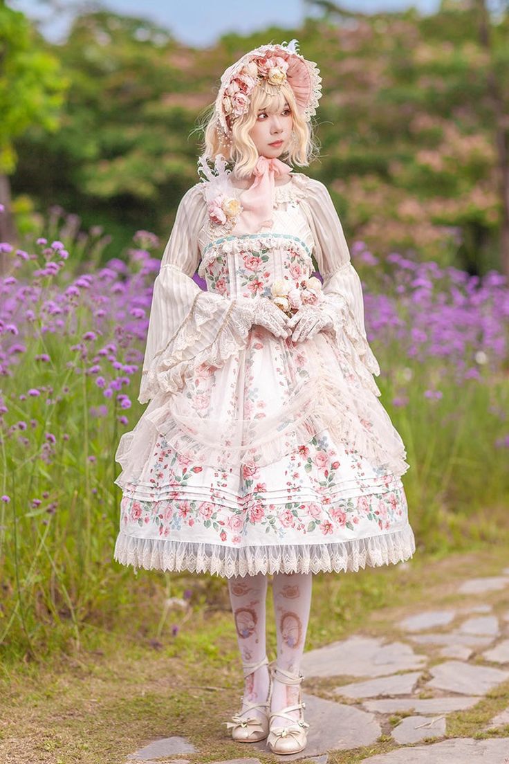 a young woman, dressed in a flowing white lolita fashion dress adorned with a delicate floral print, stands on a stone path amidst a vibrant purple flower field. she is posing for the camera, her gaze slightly turned away, showcasing the intricate details of her outfit, which includes a matching bonnet and sheer sleeves. the natural daylight illuminates the scene, highlighting the soft colors of her dress and the lush greenery surrounding her.