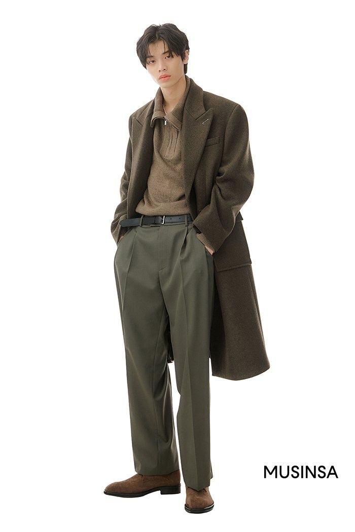 a man is standing in a studio, wearing a brown coat over a light brown sweater, with a black belt and green pants, and brown shoes. he is looking to the side with a serious expression.