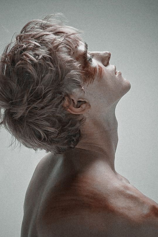 the image showcases a closeup profile portrait of a young man against a muted gray background. his head is tilted back, eyes closed, revealing a sense of serenity or perhaps vulnerability.  his short, tousled hair has a slightly unkempt appearance. he has a toned physique, and his upper body is bare. faint, reddishbrown markings resembling streaks of paint or perhaps symbolic wounds adorn his face and shoulder, adding an element of artistry and intrigue to the composition.  the overall aesthetic leans towards the dramatic and evocative, emphasizing emotion and physicality.