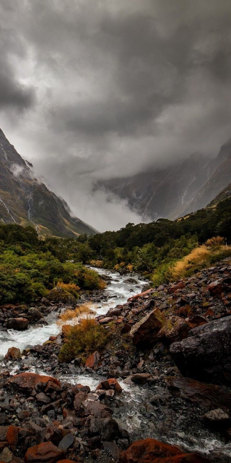 a landscape image showcasing a rocky river flowing through a mountain valley under a cloudy sky. the valley is lined with green trees and foliage, and the mountain peaks are shrouded in clouds. the river flows over rocks of various sizes, and the foreground is filled with rocks and vegetation. the overall mood of the image is dark and scenic.