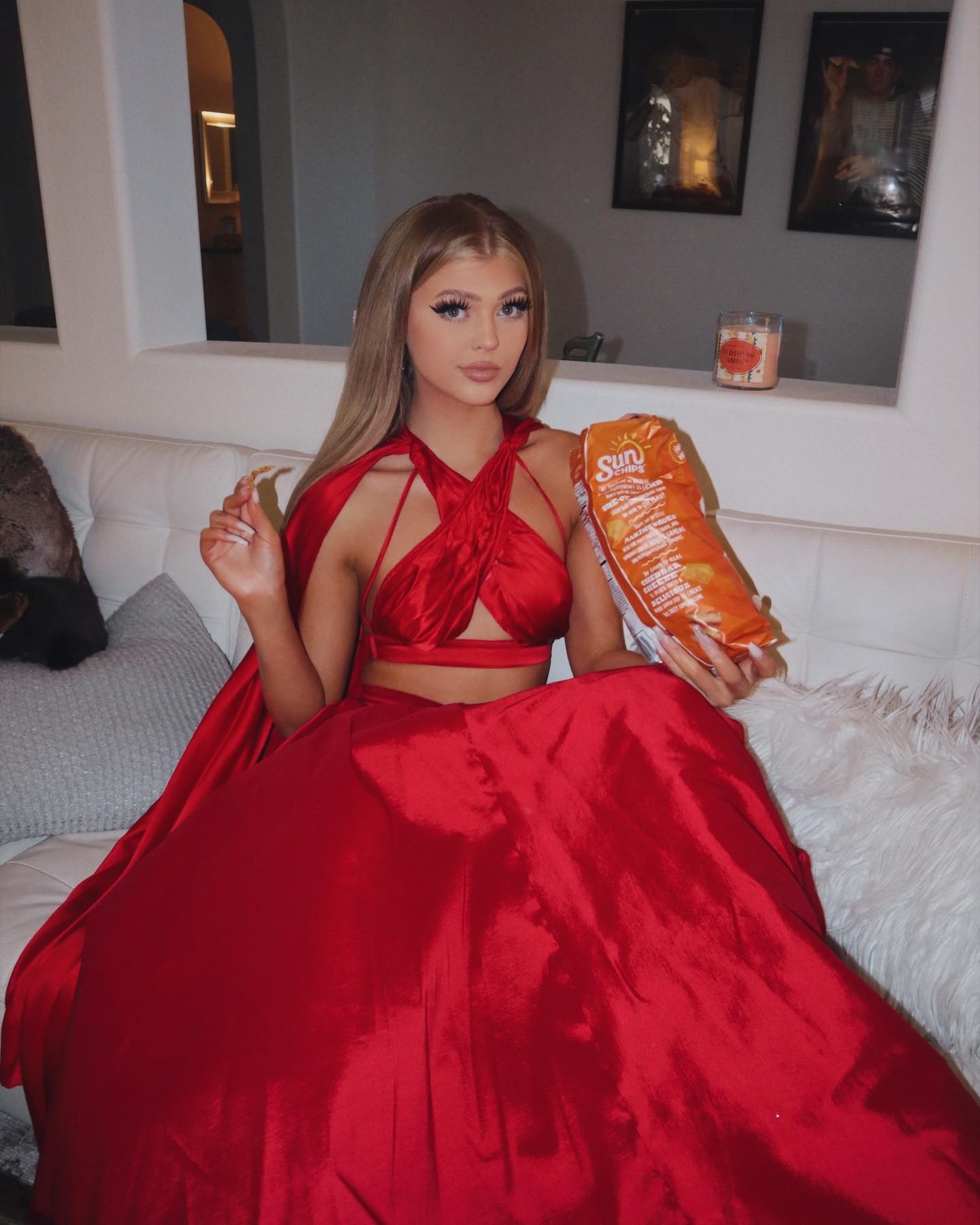 a woman with blonde hair, wearing a red two piece dress and cape is sitting on a white couch holding a bag of sun chips. there are white pillows on the couch. behind the couch are paintings. the woman has long, gold nail extensions.