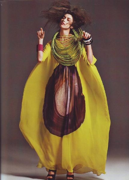 a woman in a yellow dress with a green necklace and brown patterned circle in the center stands against a grey background. she is wearing her hair down and has a slightly mischievous expression on her face. she is looking off to the side and her hands are slightly raised.