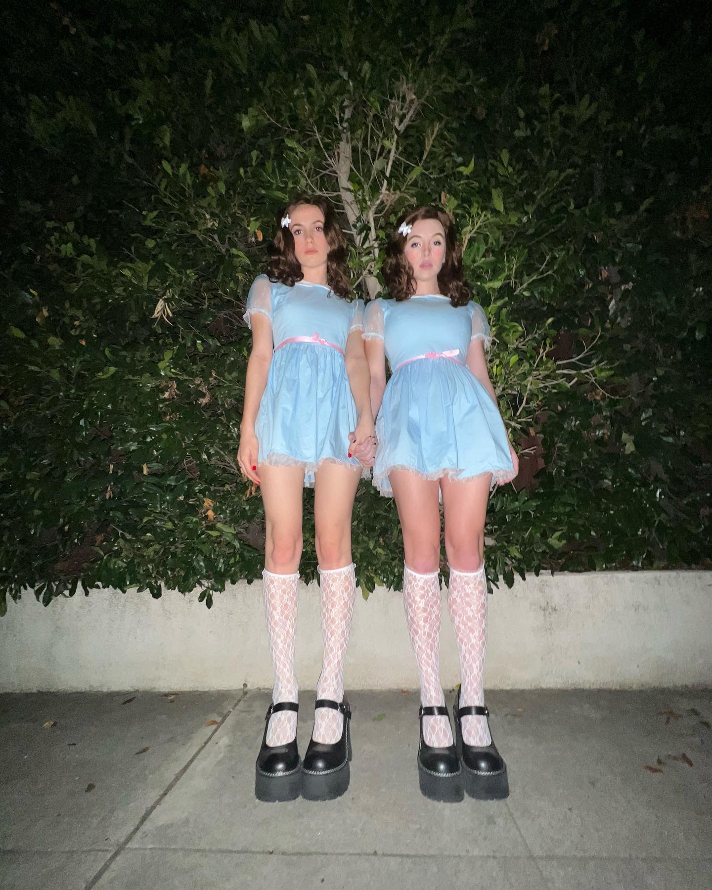 the image shows two young women dressed identically as the grady twins from stanley kubrick's film the shining. they wear pale blue dresses, white patterned kneehigh socks, and black platform mary jane shoes.  they are standing sidebyside, holding hands, in front of a dark green hedge at night. the overall aesthetic is creepy and evocative of the film's unsettling atmosphere. the costumes are meticulously replicated.