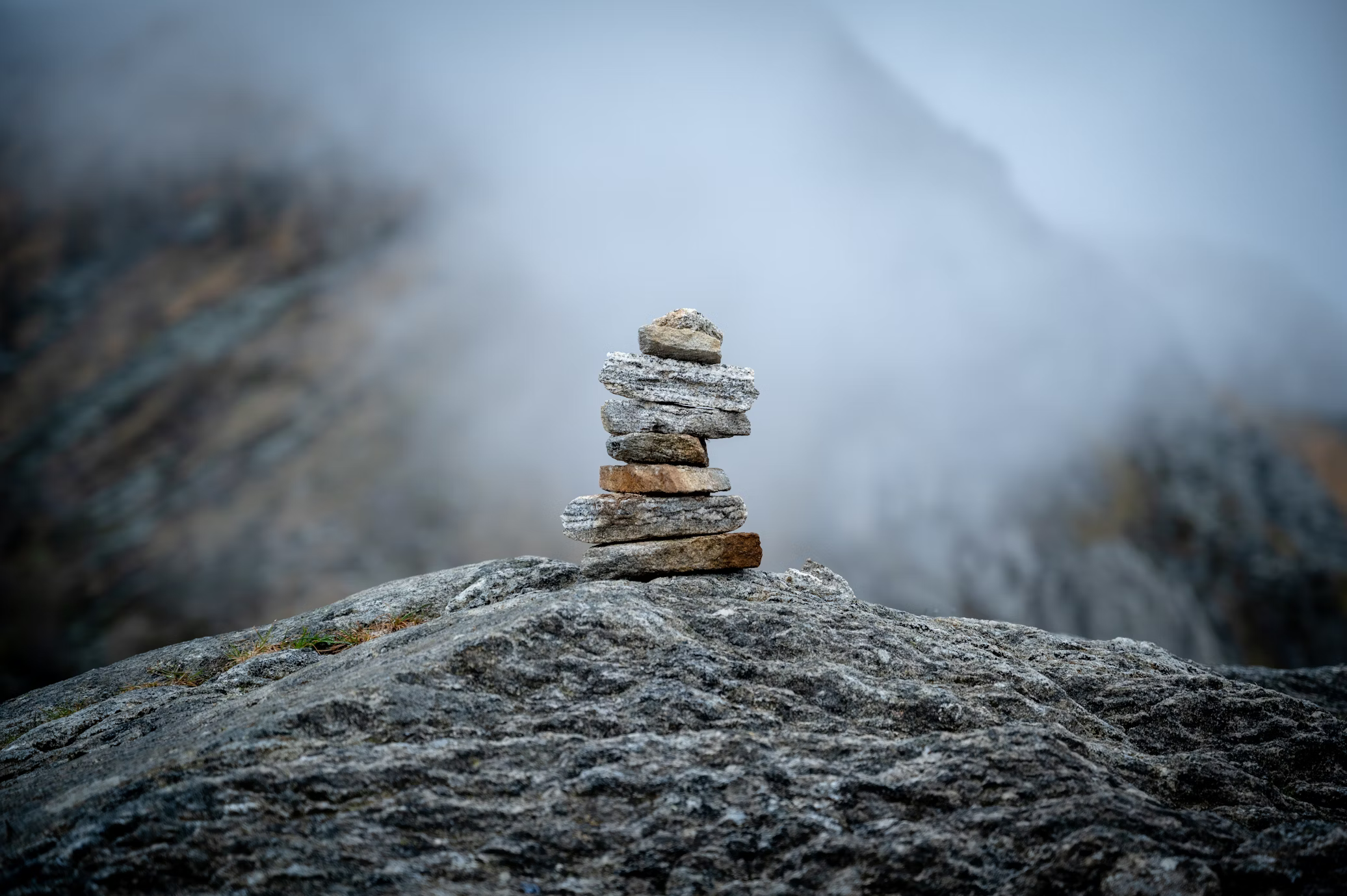a stack of rocks, known as a cairn, sits carefully balanced on a large, flat rock. the cairn is the focal point of the image, centered and sharply in focus. the surrounding environment is a mountainous landscape shrouded in a thick, white fog, giving the scene a serene and slightly mysterious atmosphere. the overall color palette is cool and muted, reflecting the overcast lighting conditions.