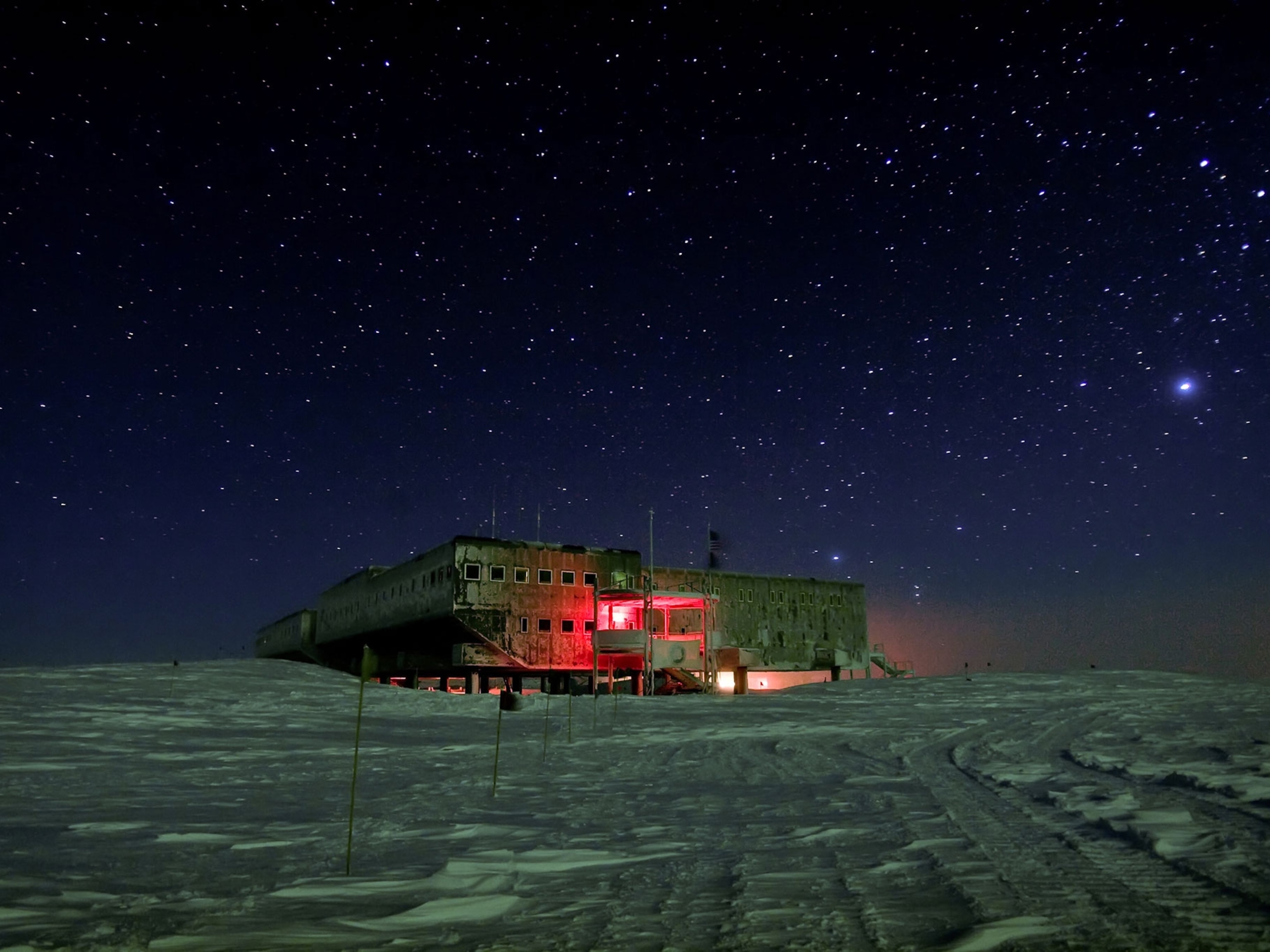 a building stands alone in a vast, snowy landscape under a sky filled with countless stars. the building is illuminated by warm, reddish light, making it appear almost inviting in the otherwise cold and desolate environment. the scene evokes a sense of isolation, mystery, and the boundless possibilities of the frozen wilderness.
