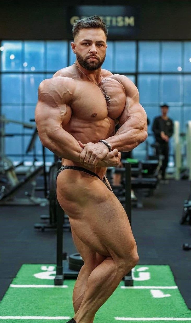 the image shows a muscular male bodybuilder posing in a gym. he is flexing his muscles, showcasing his physique. the background is slightly blurred, but shows gym equipment and another person in the background. the bodybuilder is wearing black posing trunks and a necklace. the overall tone of the image is professional, emphasizing the athlete's physical condition and accomplishments. the green mat he's standing on indicates a possible competition or training setup.