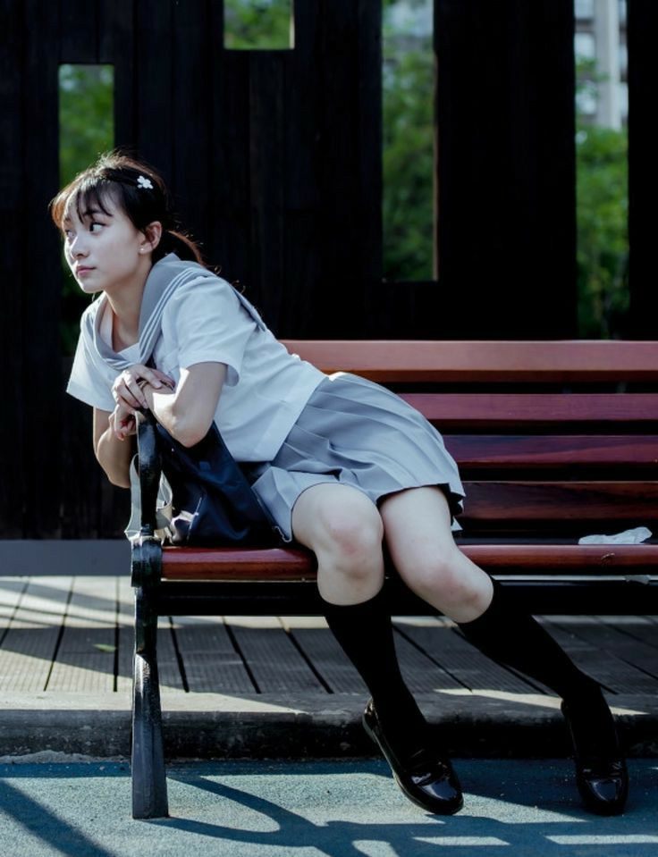 a young woman is sitting on a wooden park bench with her arms crossed. she is wearing a sailor style school uniform and dark socks and shoes. her left leg is bent with her foot resting on the bench while her right leg is straight out in front of her. she is looking at something off to the side of the photo.