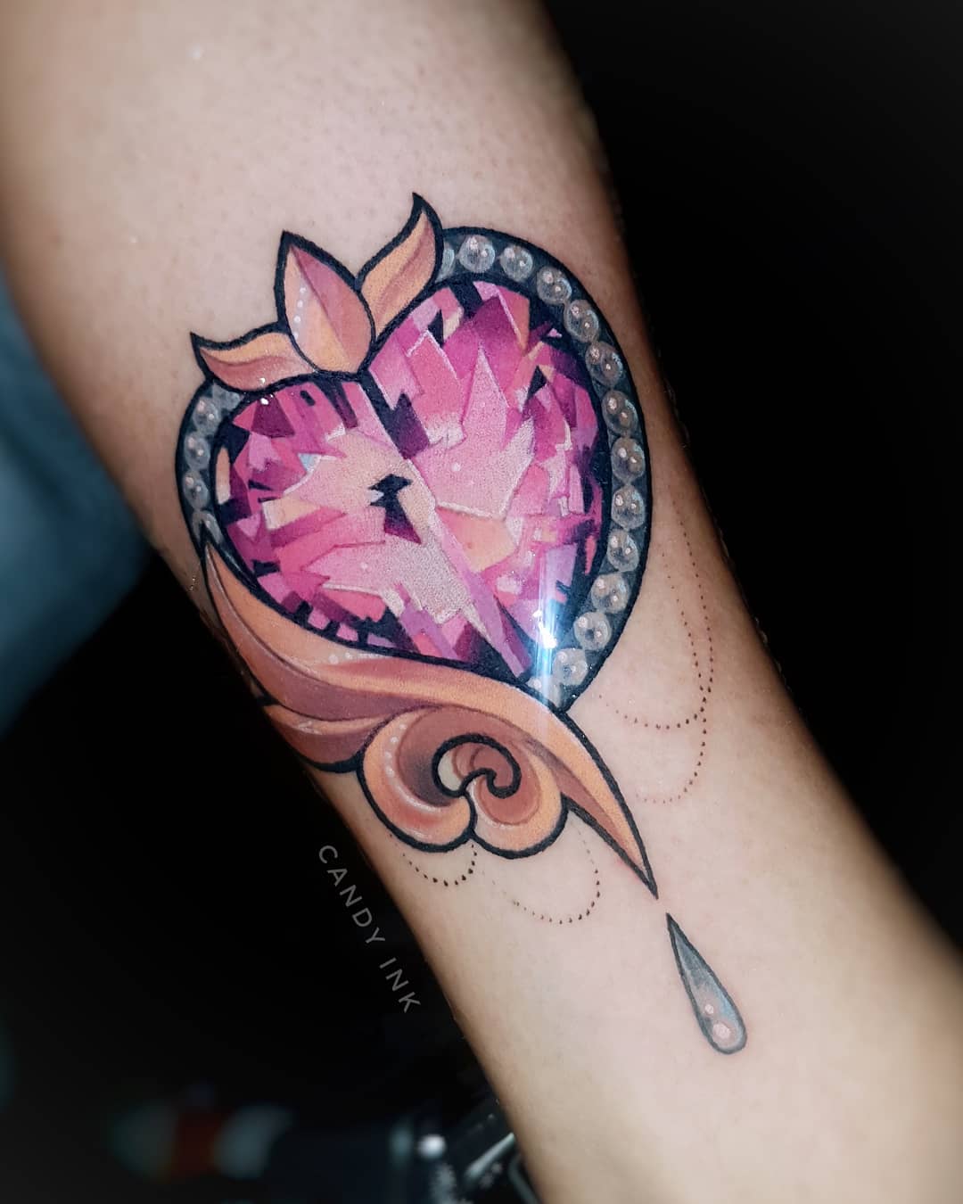 a pink jeweled heart tattoo on someone's inner arm. the heart is intricately detailed and has a drop of water falling from its point. there is some ornate scrollwork underneath the heart. the tattoo artist's signature, "candy ink," is visible below the scrollwork.