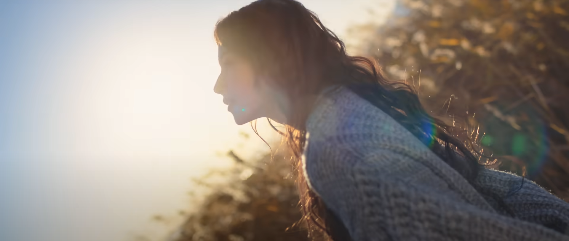 the image shows a woman in profile, her face illuminated by the setting sun. she is wearing a cozy grey sweater and has long, dark hair. the background is a field of tall grasses bathed in the golden light of the setting sun. lens flares add to the warm and ethereal feel of the image. the overall mood is one of peace, tranquility, and connection with nature.