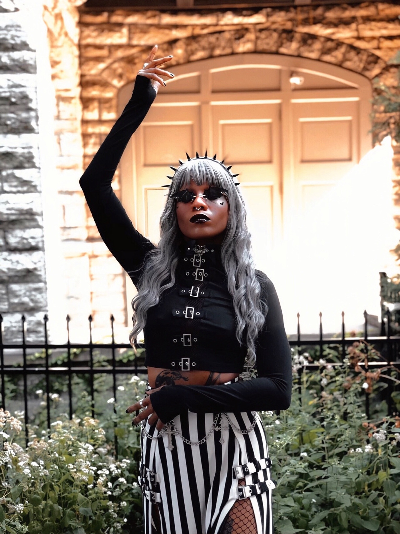 a young woman with long, silver hair and black clothing poses in front of a doorway with a metal fence in the foreground and greenery on the sides. she is wearing a black, longsleeved shirt with a silver chain and buckles, black pants with white stripes and silver buckles, and a crownlike headband with a black choker necklace. the woman is looking up and to the left. she is wearing dark sunglasses. she has tattoos on her hands.