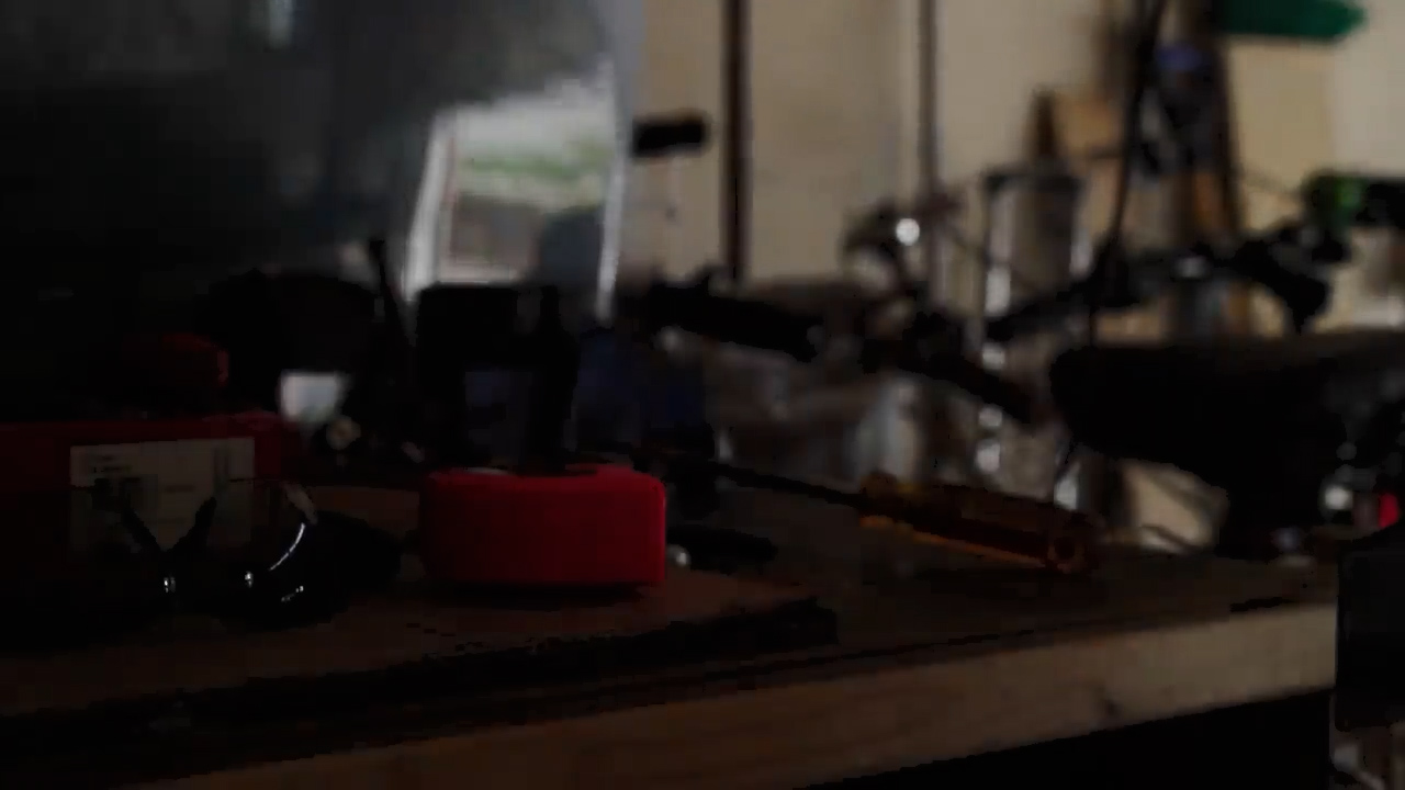 the image shows a dimly lit workbench in a garage. there are various tools on the workbench, including a screwdriver, safety glasses, and a red tape measure. a bicycle is visible in the background, partially obscured by the darkness.  the overall impression is of a workspace used for repairs or maintenance.