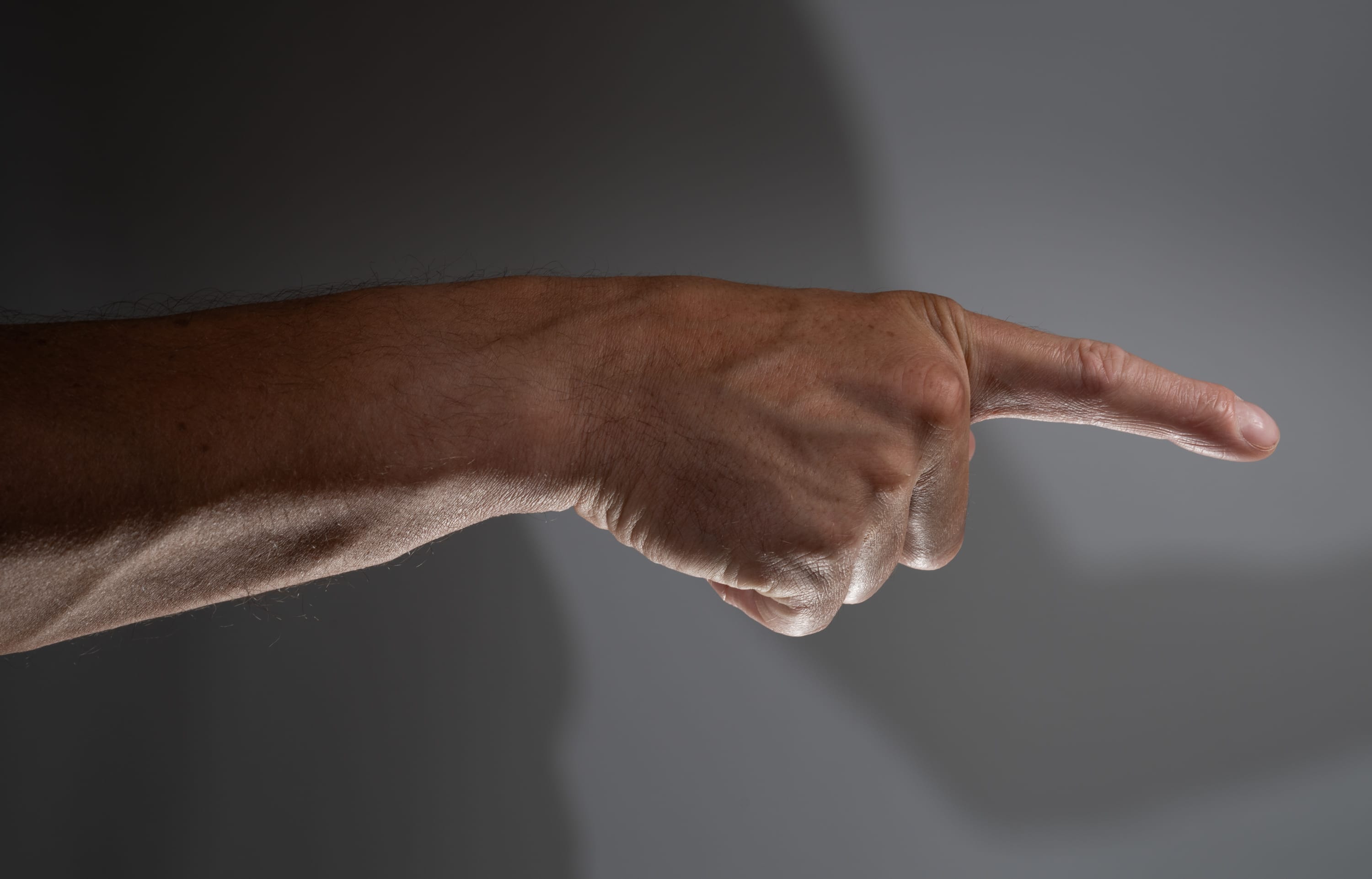 a man's hand is extended against a gray background, his index finger pointing towards the right side of the frame. the hand is illuminated with studio lighting, casting a shadow on the background.