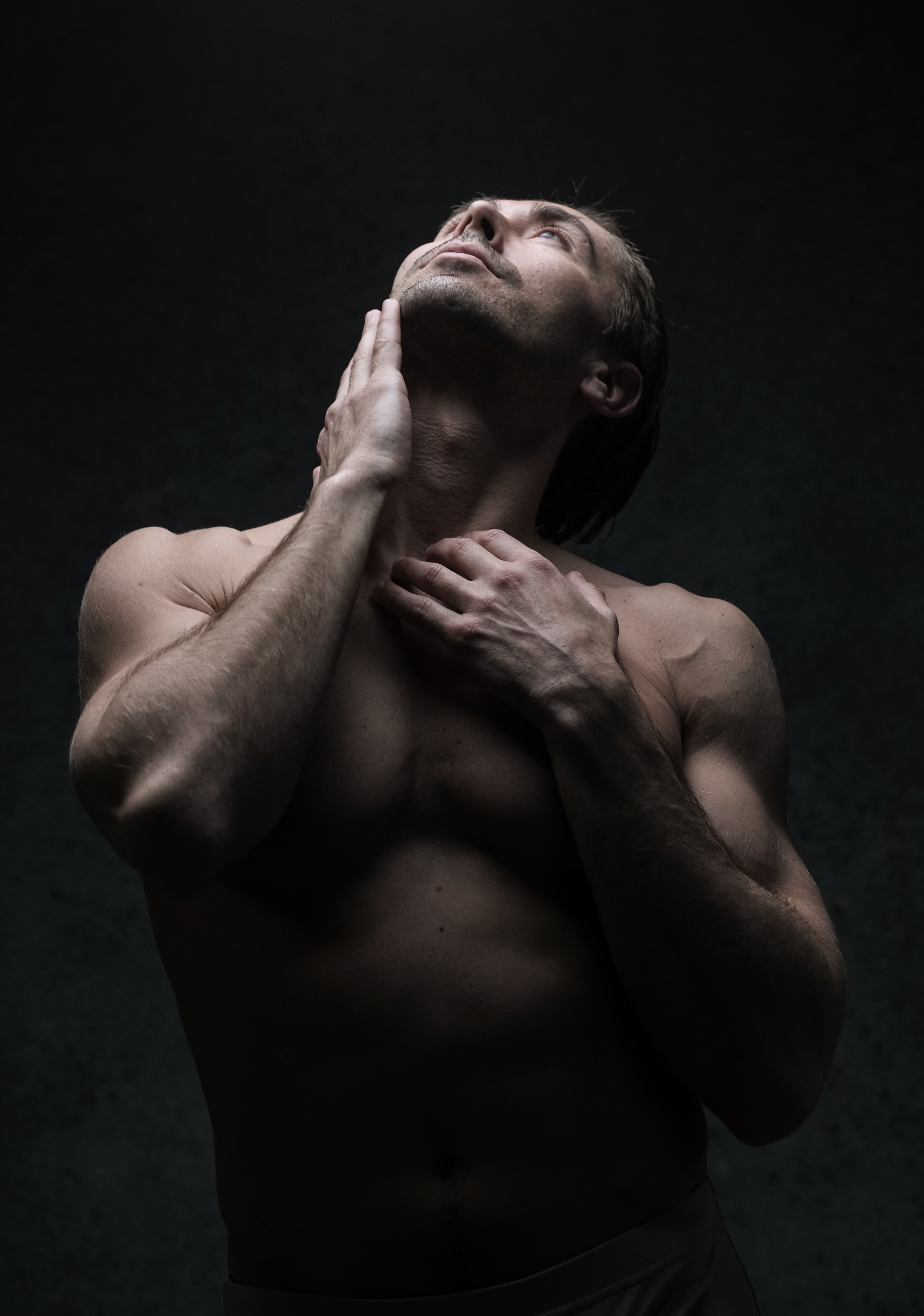 a muscular man is shown from the waist up against a dark background. he is shirtless and looking upward, his hands gently touching his neck. the lighting is dramatic, creating deep shadows and highlighting his physique.