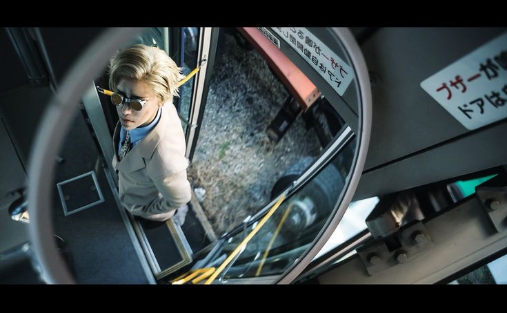 the image is a stylish, highangle closeup shot of a fashionably dressed blonde man seen through a bus's interior mirror. he's wearing a light beige suit, a light blue collared shirt, and distinctive goggles. the bus's interior is visible, showing some mechanical details and japanese text on signage. the overall aesthetic is modern and urban. the reflection in the mirror adds depth and intrigue to the photograph, creating a stylish and slightly surreal feel.
