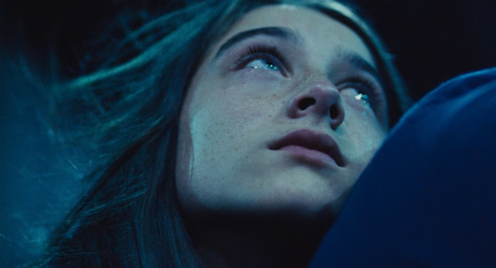 a young woman with long dark hair and a pale complexion looks upwards with a sad expression on her face. she is closeup, and her eyes are welled with tears. the scene is lit with a blue light that casts shadows over the woman's face. the image is dark and moody and portrays an emotion of sadness and vulnerability.