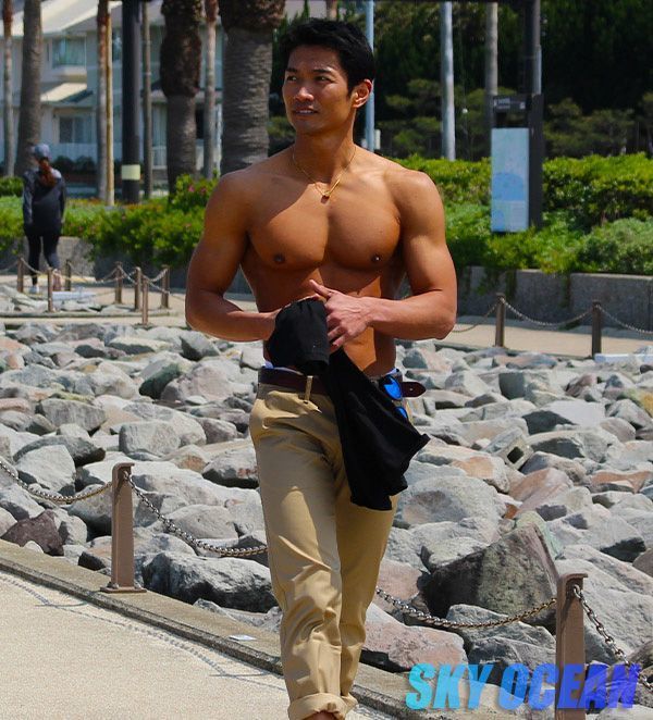 a shirtless, muscular asian man with a toned physique walks along a path lined with rocks near the ocean. he is holding a darkcolored shirt in his hands.  he is wearing khaki pants and has a relaxed yet stylish look. the background shows a coastal scene with palm trees and buildings. the overall image is bright and sunny, suggesting a summer day.