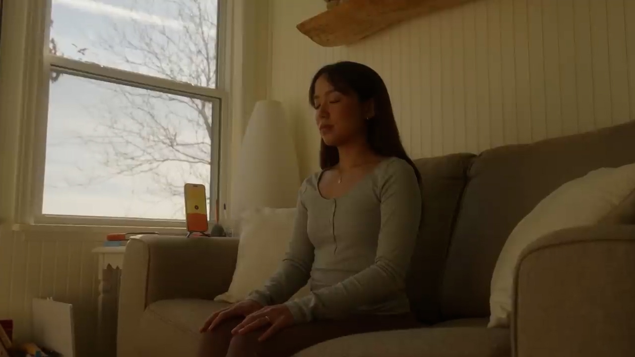 a young woman sits on a couch with her eyes closed, appearing to be meditating. there's a window behind her with a view of a tree and blue sky. soft, natural sunlight illuminates the room, casting warm shadows. a phone rests on a table in front of the window. the overall atmosphere is one of peace and tranquility.