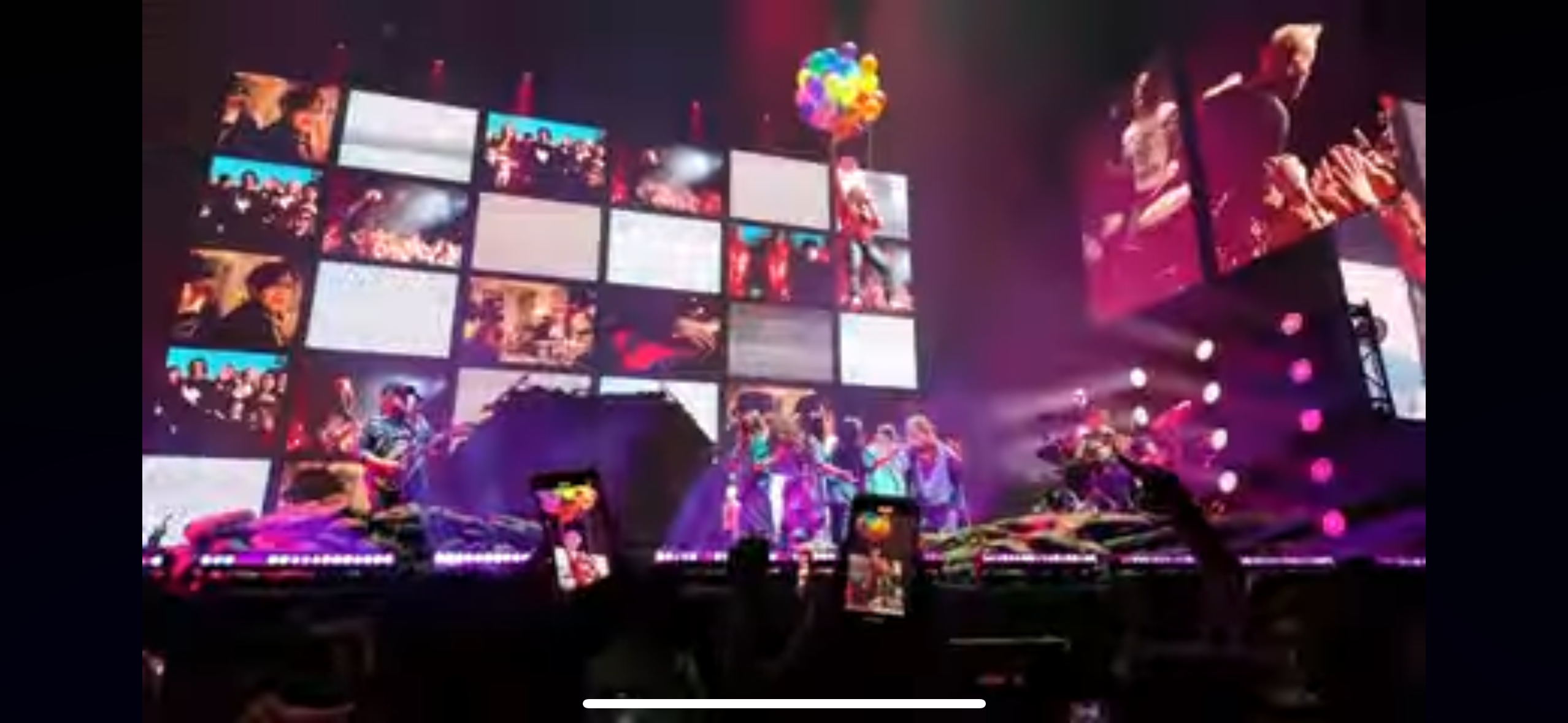 a live music concert is taking place, with a band performing on stage in front of a large crowd. the stage is brightly lit and there are large video screens displaying images of the band and the crowd. the band members are playing instruments and singing to a lively crowd. the atmosphere is electric, with the music, lights, and energy creating a truly memorable experience.