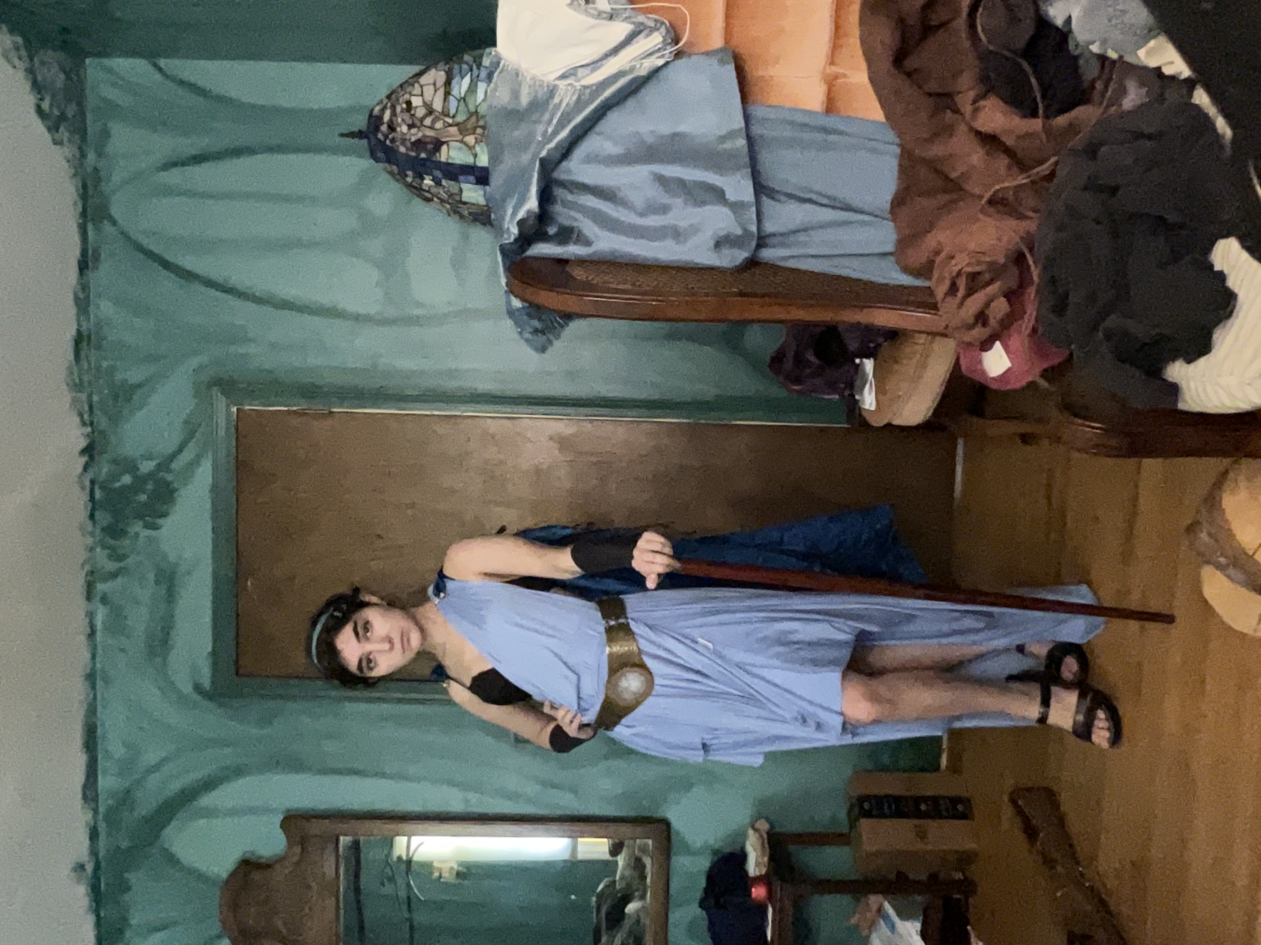 a person is cosplaying in a blue togastyle dress.  they are standing in a room with teal walls and a wooden floor. the room is somewhat cluttered with clothing and other items. the cosplayer is holding a cane and wearing sandals.  they have on a headband and appear to be a woman.  a partially visible mirror and chair are in the background.