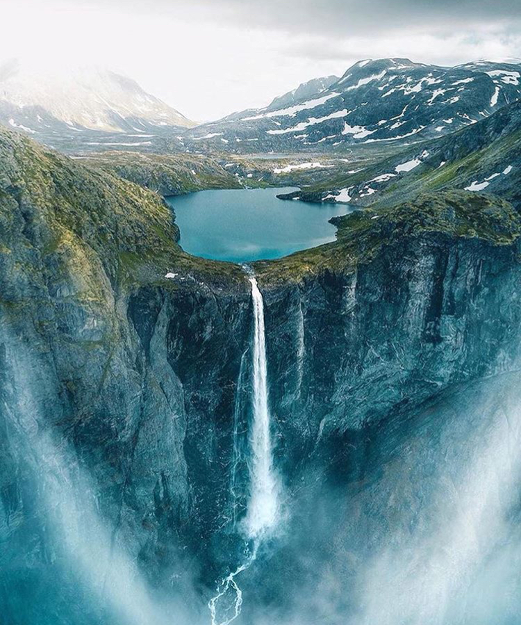 the image is a highangle shot of a waterfall cascading from a lake atop a cliff. the surrounding area consists of mountains and a valley, with some snow visible on the mountain peaks. the sky is cloudy, and the overall atmosphere is one of natural beauty and grandeur.