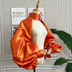 a closeup image of a vibrant orange silk jacket, casually draped over a mannequin. the jacket boasts a unique design with voluminous, gathered sleeves and a high collar, adding a touch of drama and sophistication.  the background features a framed paristhemed print and a glimpse of lush greenery.