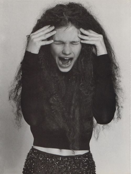 a black and white image of a young woman with long, curly hair. she is wearing a black sweater and a black sequined skirt and is screaming with her eyes closed and her hands on her head. she looks distressed and emotional. the image is lit from the front and there is a white background.