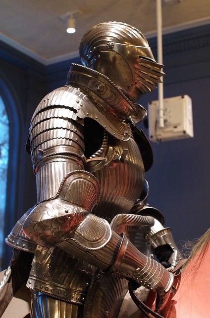 a closeup image of an intricately crafted suit of plate armor displayed on a mannequin inside a museum. the armor is made of polished metal with a series of ridges and fluting, likely meant to deflect blows. the helmet, with its visor raised, features a distinctive design with a series of vertical slits. the low angle shot emphasizes the imposing nature of the armor. the lighting is artificial, likely from spotlights, highlighting the details and craftsmanship of the piece.