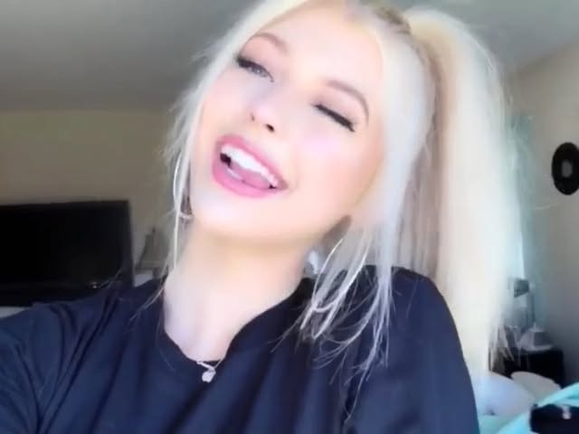 a woman with blonde hair pulled into pigtails is wearing a black shirt, earrings, and a necklace. she is sticking her tongue out and winking. the background appears to be a bedroom.