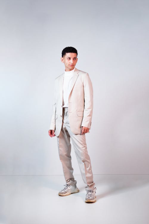 the image shows a young male model standing against a light grey background. he is wearing a complete beige suit consisting of a blazer and trousers. underneath, he sports a creamcolored turtleneck. his shoes are light grey sneakers. the model is positioned in a slightly turned pose, creating a dynamic feel. the overall aesthetic is minimalist and sophisticated, showcasing the outfit's contemporary style and neutral color palette. the setting is a clean studio environment with even lighting, highlighting the outfit details and the model's pose.