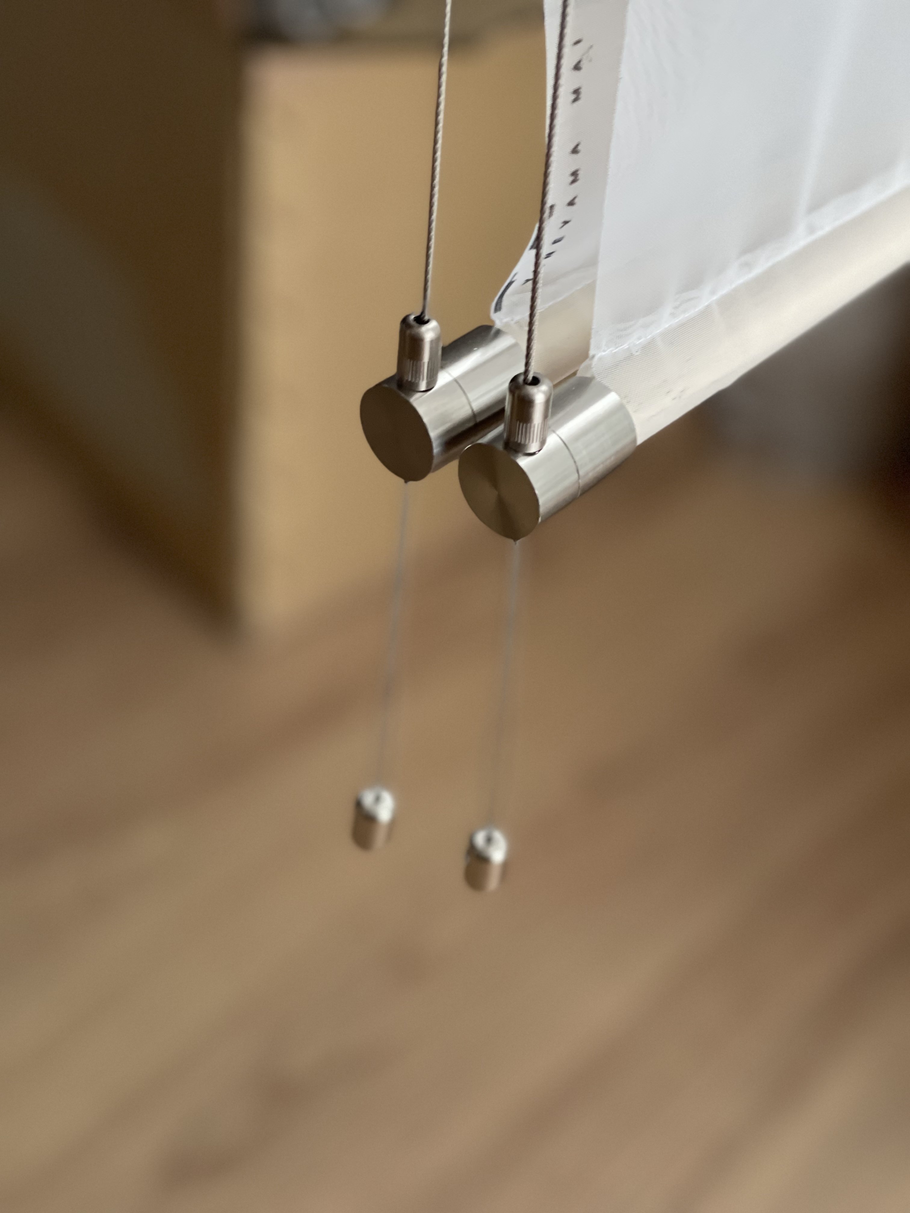 the image shows a closeup view of a modern curtain rod made of metal. the rod is silver and has a minimalist design. it is attached to the wall with two cables and has a white curtain hanging from it. the background is blurred and shows a wooden floor and a beige wall.