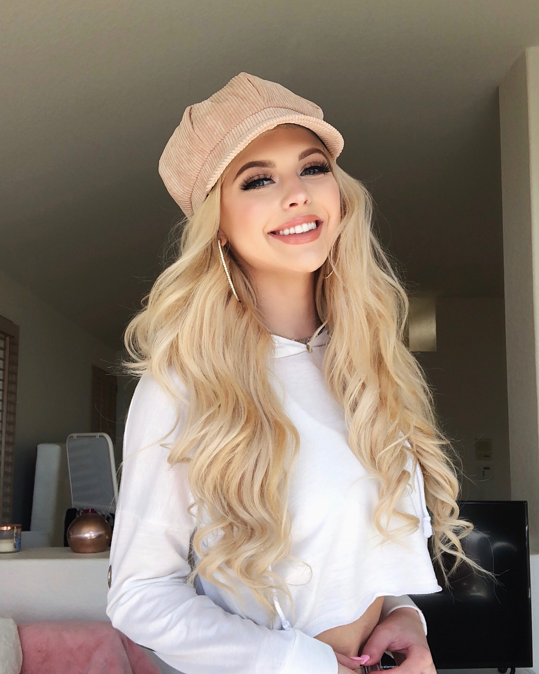 a woman with long blonde hair, wearing a light pink corduroy newsboy hat, a white crop top, and gold hoop earrings, smiles into the camera while posing indoors. behind her is a room with a television, a lit candle, and other decor.
