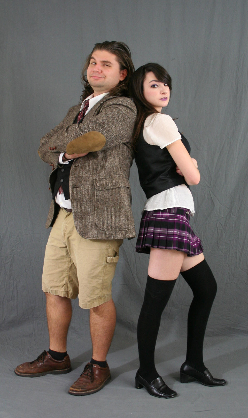 a full shot of a man and woman standing backtoback against a gray background. the man is wearing a tweed jacket, khaki shorts, and loafers. he has his arms crossed and a slight smirk on his face. the woman is wearing a white shirt, black vest, plaid skirt, and black thighhigh socks and loafers. she is posing with her arms crossed and a slightly serious expression. the image is playful and suggestive of a cosplay or costume scenario.