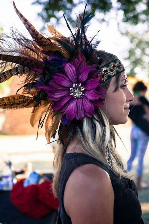 a young woman with blonde hair is wearing a purple flower headdress made of feathers and a headband. she is smiling and looking to the right. the woman is outdoors, surrounded by summer greenery.