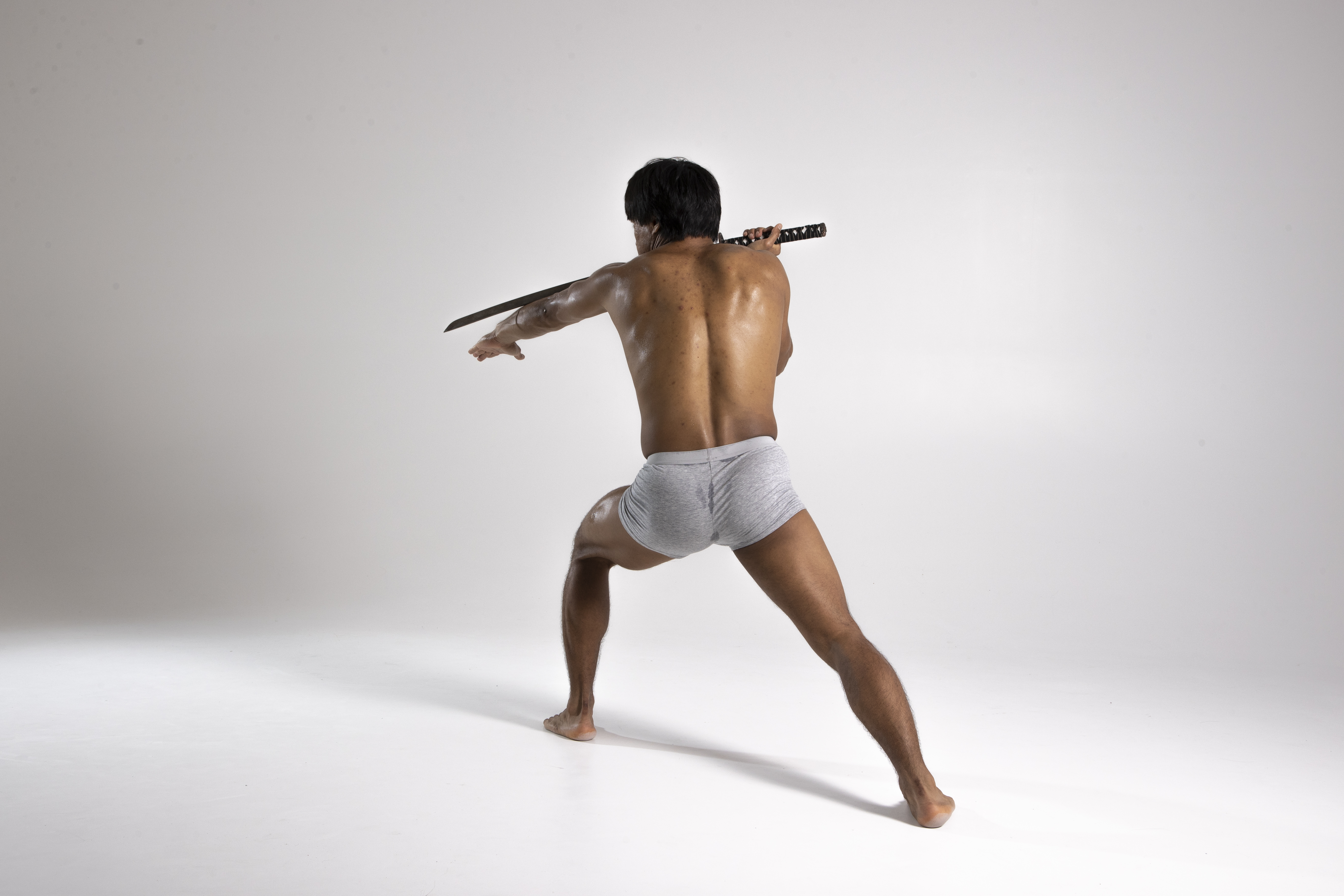 a shirtless man wearing underwear is seen from the rear view, holding a sword indoors against a white background. he appears to be in a warrior stance.