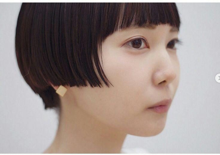 a young woman with short, black, bluntcut bob hair and bangs is looking to the right with a serious expression. she is wearing a pair of gold squareshaped earrings. the background is white.