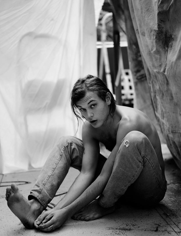 a young man, shirtless and wearing jeans, sits on the ground looking directly at the camera. the background is blurred and the photo is in black and white.