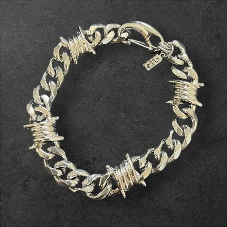 a closeup shot of a silver barbed wire chain bracelet. the bracelet has a curb chain style and is made of metal with barbed wire details. the clasp is visible in the top right corner of the image. the bracelet is set against a black background.