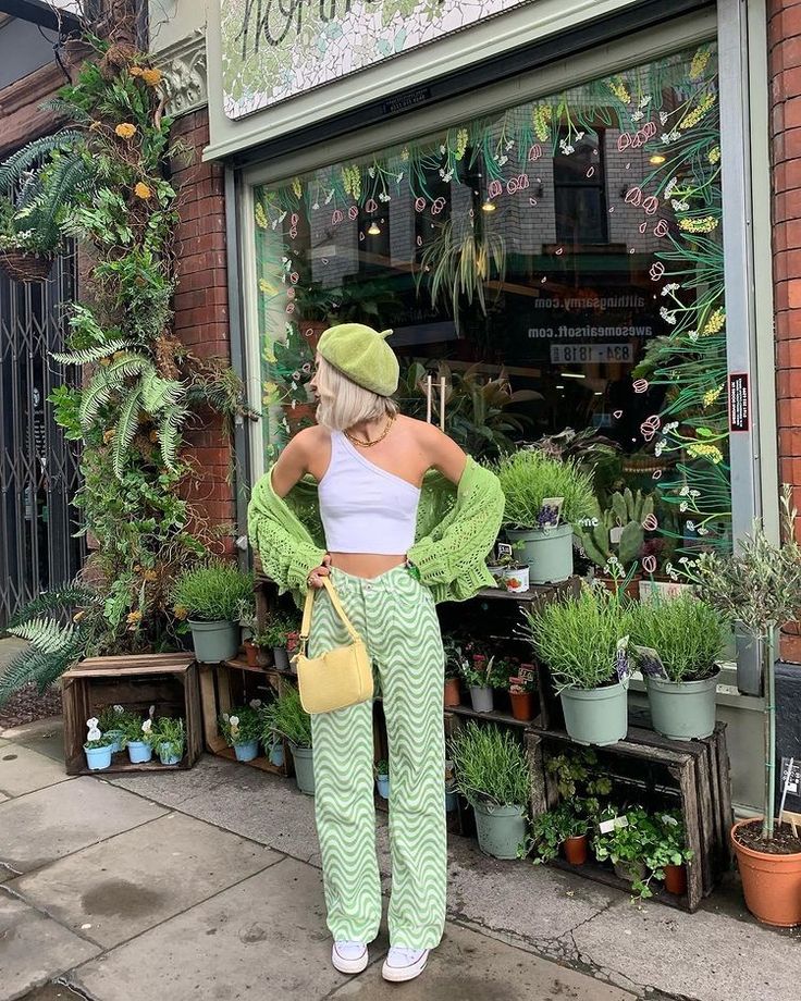 a young woman in a green crochet cardigan, white tank top, and green patterned pants with a yellow bag poses outside a shop with greenery and flowers in the window. she's wearing a green beret and is looking to the right of the frame.