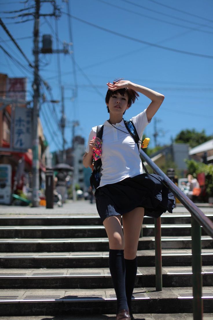 a young woman in a school uniform walks up a flight of stairs on a sunny day. she is wearing a white shirt, black skirt, black socks and shoes, and has a backpack slung over one shoulder. she is shielding her eyes from the sun with her hand. the image is taken from a low angle, giving the impression of looking up at the woman. the background is a typical japanese city street scene, with buildings, power lines, and trees visible. the overall impression is one of summer heat and youthful energy.