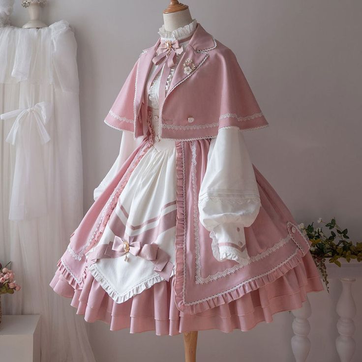 a pink and white lolita fashion dress is displayed on a mannequin. the dress features a capelet, lace trim, bows, and ruffles. the background is a white wall with a white curtain and a vase of flowers. the lighting is bright and natural.