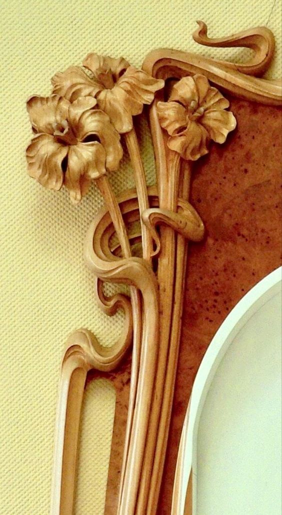 the image shows a closeup detail of a beautifully carved wooden piece, likely from a piece of furniture or architectural element. the style is clearly art nouveau, characterized by its organic, flowing lines and floral motifs.  three stylized irises are prominently featured, carved with great detail and realism. the wood is a light, golden hue, showing visible grain. the carving is intricate, with swirling lines and elegant curves that interweave with the floral elements. the overall effect is one of exquisite craftsmanship and refined aesthetic beauty. the background is a pale yellowgold, which contrasts nicely with the warm tones of the wood.