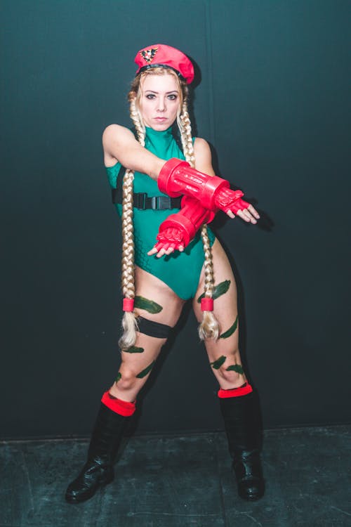 a full shot of a woman cosplaying as cammy from the street fighter video game series. she is wearing a green bodysuit, red beret, red gloves, and black boots. her blonde hair is styled in pigtails, and she has green camouflagelike markings painted on her legs. she is striking a fighting pose, and the background is dark.