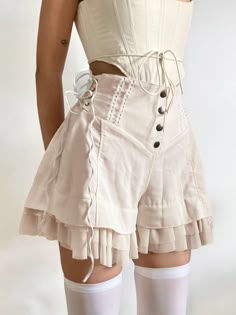 a woman is wearing a white corset and white skirt with tulle ruffles. she is also wearing white thighhigh stockings and has a white backdrop.