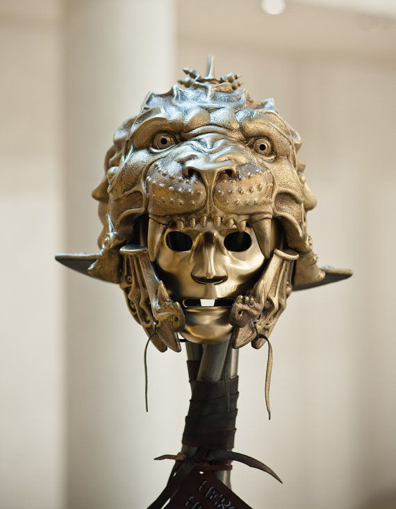 a closeup shot features a goldenbronze metal sculpture of a helmet, resembling a lion's head with a humanlike face underneath. the lion's face is detailed with eyes, nose, and teeth, while the lower face has a smooth, expressionless appearance. the helmet is mounted on a dark, wrapped stand, and the background is a blurred, neutral tone.