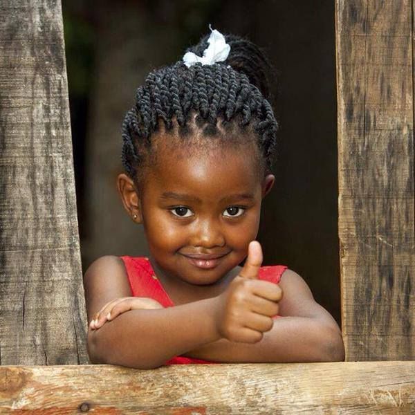a young african girl with braided hair and a white hair bow is shown from the chest up, giving a thumbs up while leaning on a wooden frame. she is wearing a red dress and has a slight smile on her face. the background is dark and out of focus.