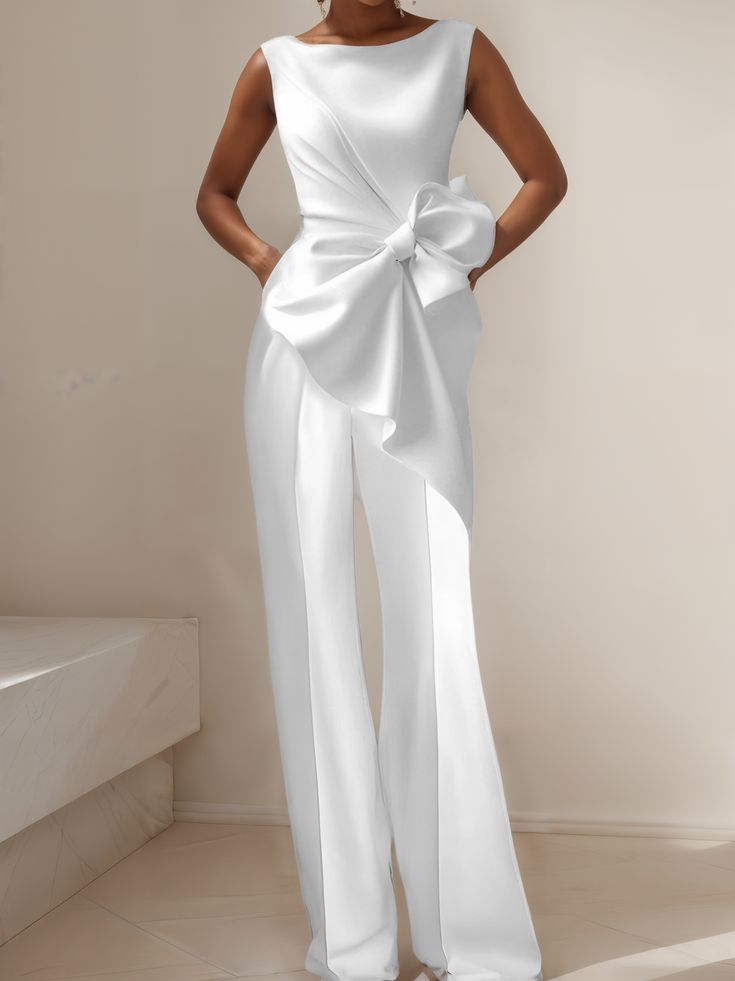 a woman wearing a white jumpsuit with a large bow at the waist. she is posing in a studio with a white background and a white wall behind her. the jumpsuit is very elegant and sophisticated, and the model looks very chic and fashionable.