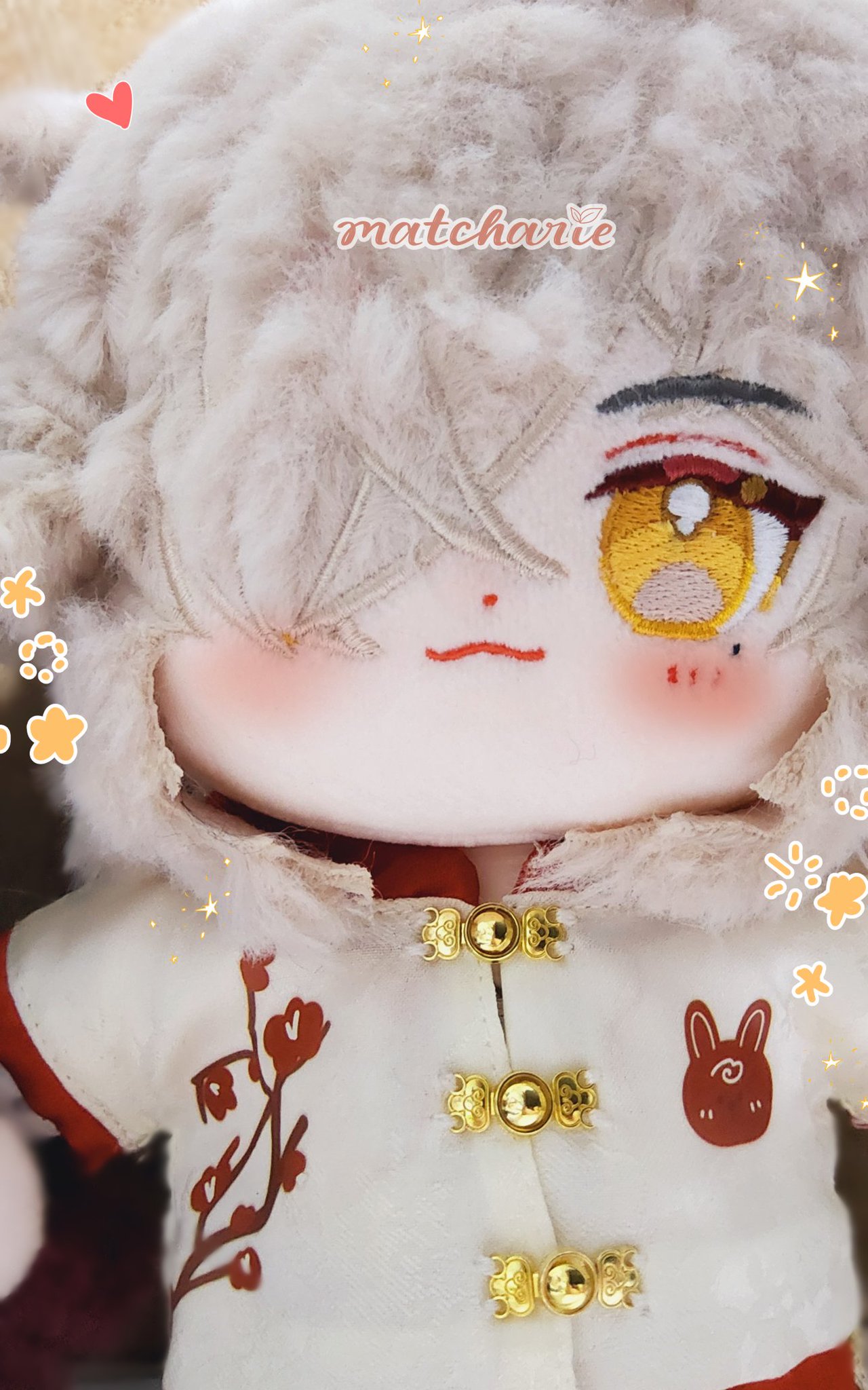 a plush doll with white fur,  red eyes and a cute expression is dressed in white with red embroidered flowers and golden buttons. the doll is facing forward and the focus is on its face and clothes.