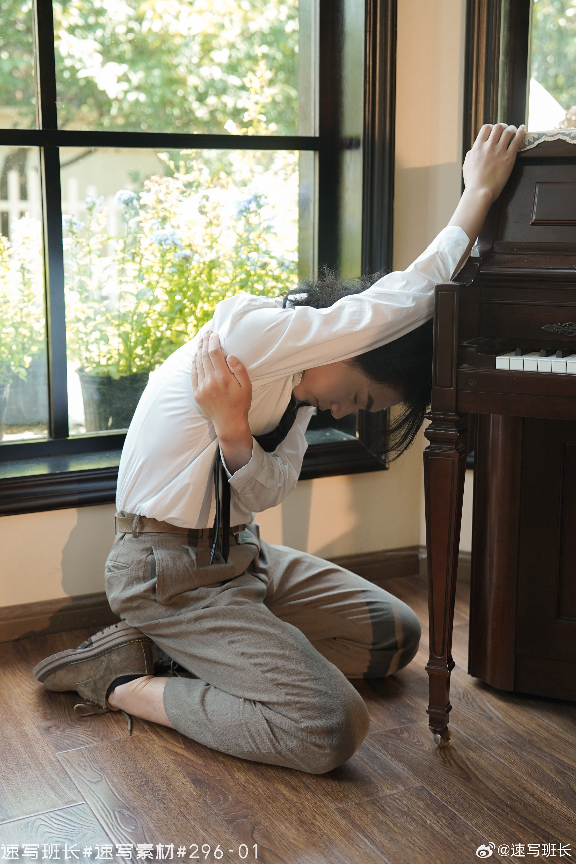 a man with long dark hair is kneeling in front of an upright piano, with his back to the viewer. his arms are stretched upwards, reaching for the piano. he is wearing a white shirt, brown pants, and brown shoes. sunlight is streaming through a window behind him, illuminating the scene. the overall mood is somber and contemplative.