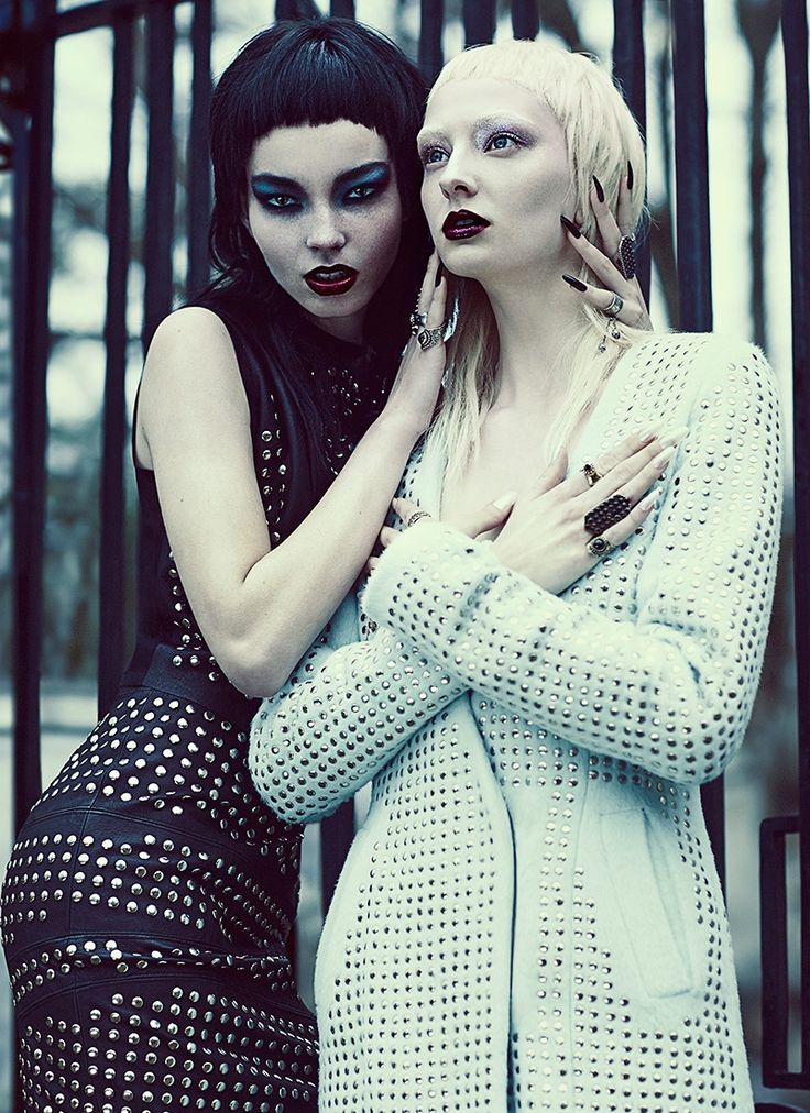 two women pose in fashion attire against a metal fence background. the woman on the left is wearing a black studded mini dress and has black hair with bangs. the woman on the right is wearing a white studded cardigan with blonde hair. the woman on the left is looking at the camera while the woman on the right is looking away.  both women are wearing several pieces of jewelry.