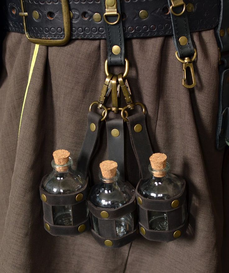closeup image showing three small glass potion bottles, each fitted with a cork stopper and enclosed in a black leather pouch, hanging from a black leather belt with metal fasteners. the belt is decorated with multiple brasscolored rings and has a brass buckle. the bottles are suspended via metal clasps attached to leather straps. the background is a plain brown fabric.